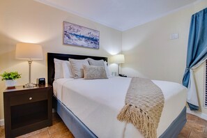 A neatly made bed with a white comforter and a beige throw blanket, flanked by two bedside tables with lamps, a wall-mounted painting, and a window with curtains.
