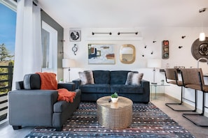 Een moderne woonkamer met een grijze bank, een blauwe bank en een ronde salontafel. Er staan twee lampen, een klok en een wandklok.