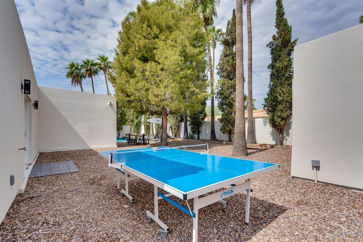 A ping pong table in an outdoor area with gravel flooring, surrounded by trees and white walls.