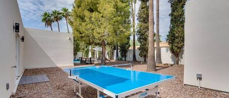 A ping pong table in an outdoor area with gravel flooring, surrounded by trees and white walls.