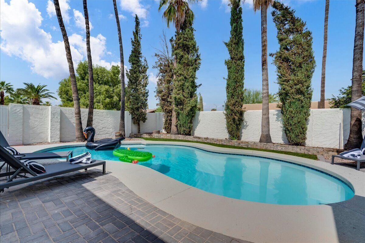 A swimming pool with lounge chairs, surrounded by tall trees and a white wall.