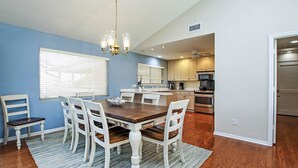 A dining area with a wooden table and white chairs, a chandelier, and a kitchen in the background. - Sandcastle Home on Sanibel Island in The Dunes Golf Club (Sanibel)