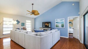 A spacious living room with a large gray sectional sofa, a ceiling fan, a flat-screen TV, and a painting on the wall. - Sandcastle Home on Sanibel Island in The Dunes Golf Club (Sanibel)