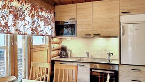 A kitchen with wooden cabinets, a refrigerator, a coffee maker, and a window with autumn leaves. - Salmikankaan kelo d2 by Interhome (Kuusamo)