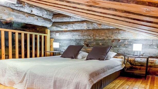 A bedroom with a wooden ceiling, a bed with a white bedspread and two pillows, a wooden floor, and a small table with a lamp. - Salmikankaan kelo d2 by Interhome (Kuusamo)