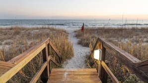 Eine Holzpromenade, die zu einem Strand führt, im Hintergrund ist ein Rettungsturm sichtbar.