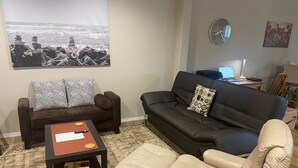Una sala de estar con un sofá marrón, un sofá negro y un sillón beige. Hay una mesita con un libro y un control remoto. Una alfombra está en el suelo y un cuadro en la pared.