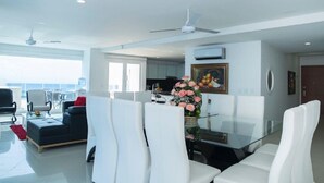 A modern living room with a glass dining table, white chairs, a dark sofa, and a red accent rug. There are ceiling fans, a kitchen in the background, and a large window offering a view. - PH17P Penthouse Cartagena Frente al mar (Cartagena)