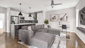 Een moderne woonkamer met een grijze modulaire bank, een keuken met witte kasten, een eethoek met een witte tafel en kunstwerken aan de muren.