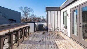 Een terras met houten vloer, metalen barstoelen en een glazen deur die naar een kamer leidt.