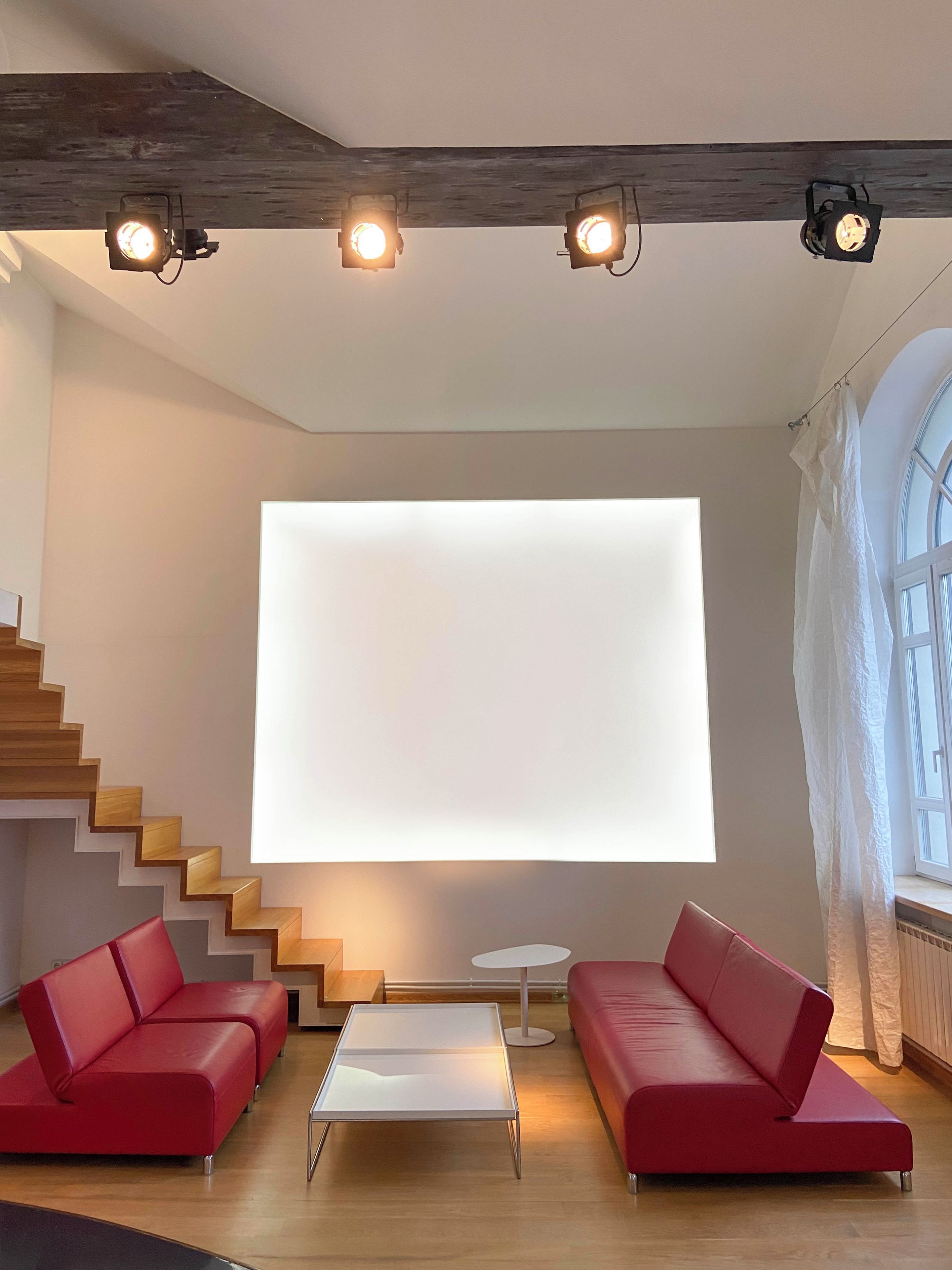 A modern living room with a staircase, red sofas, a glass coffee table, and a large white screen.