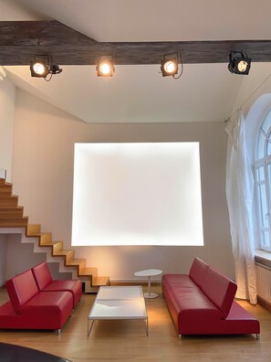 Una sala de estar moderna con una escalera, sofás rojos, una mesa de centro de cristal y una pantalla blanca grande.
