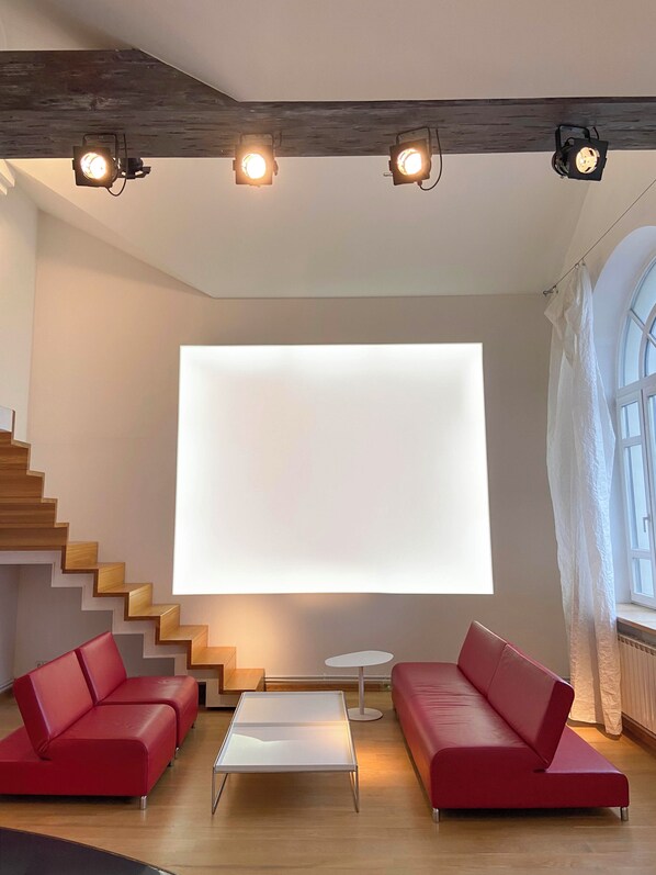 A modern living room with a staircase, red sofas, a glass coffee table, and a large white screen.