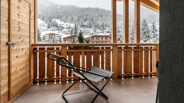 A wooden balcony with a folding chair, wooden railings, and a view of snow-covered buildings and trees.