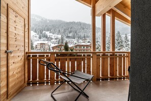 Un balcón de madera con una silla plegable, barandillas de madera y vistas a edificios y árboles cubiertos de nieve.
