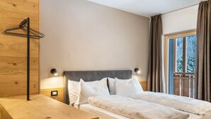 Une chambre à coucher avec une table de chevet en bois, un lit avec une literie blanche, une tête de lit dotée d’un éclairage intégré et une fenêtre avec des rideaux.