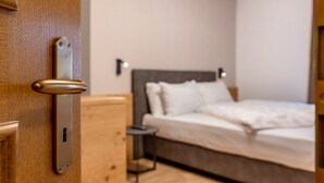 A wooden door with a modern lock, a bed with white bedding, a bedside table, and a wall-mounted light.