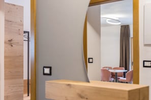 A modern hotel room with a wooden accent wall, a round mirror, a small table, and a dining area with pink chairs. - Kamerloy Apartments - D with Mountain View, Wi-Fi, Balcony and Garden (Campitello di Fassa)