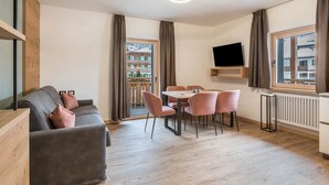 Un salon moderne comprenant un canapé, une table à manger, des chaises, un téléviseur fixé au mur et une vue sur un immeuble par la fenêtre.