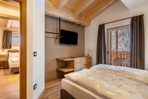 Une chambre à coucher avec un lit, un téléviseur fixé au mur, un bureau en bois et une fenêtre avec des rideaux.