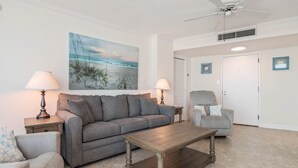 Ein Wohnzimmer mit grauem Sofa, einem Holztisch, zwei Nachttischen mit Lampen, einem Deckenventilator und einem Gemälde von einer Strandansicht.