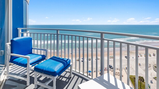 Ein Balkon mit zwei blauen Sitzgelegenheiten, einem weißen Geländer und Blick auf Strand und Meer.