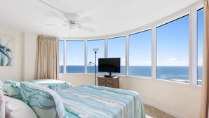 Ein Schlafzimmer mit einem großen Erkerfenster, einem Deckenventilator, einem Flachbildfernseher und einem Bett mit gestreifter Bettwäsche.