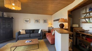 A cozy living room with a sofa, a coffee table, and a lamp. There is a kitchen area with shelves and a clock. - Résidence de l'Oisans 4 rooms - Quartier Cognet (Alpe d'Huez)