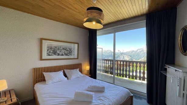 A hotel room with a large bed, a wooden ceiling, a framed picture on the wall, and a balcony with a mountain view. - Résidence de l'Oisans 4 rooms - Quartier Cognet (Alpe d'Huez)
