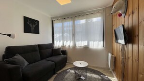 A living room with a black sofa, a small round table, a mounted TV, and a window with sheer curtains. - ASH - Cosy studio - La Menandière (Alpe d'Huez)