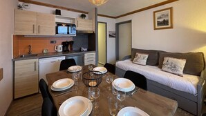 Eine kompakte Küche mit modernen Geräten, ein Esstisch für vier Personen und eine gemütliche Wohnbereits mit Sofa und Kissen.