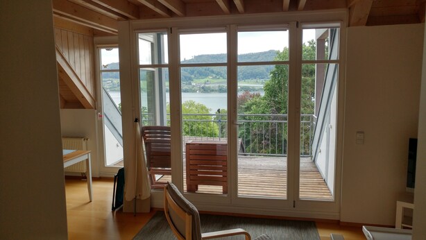 A room with a large sliding glass door leading to a balcony with a view of a lake and hills. - Vacation apartment for 3 on a hillside with a dream view of the lake (Öhningen)
