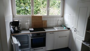 A compact kitchen with white cabinets, a built-in oven, a microwave, a coffee maker, and a window with a view of greenery. - Vacation apartment for 3 on a hillside with a dream view of the lake (Öhningen)