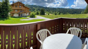A wooden deck with white plastic chairs and a table, overlooking a mountain landscape and a chalet-style building. - 3-room Apt Les Saisies, 6 ppl, Pets allowed (Les Saisies)