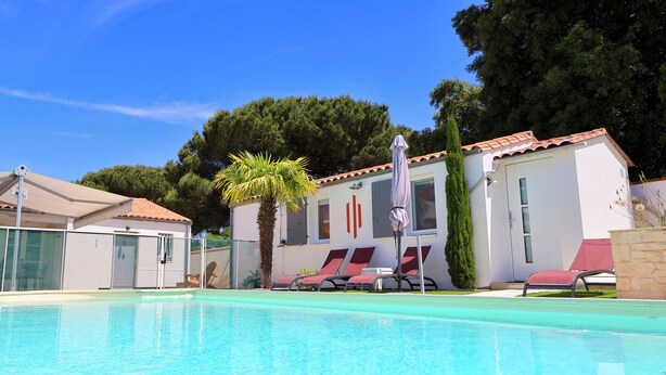 Zona de piscina con una piscina azul cristalina, sillas de descanso rojas y un edificio blanco con tejado de azulejos.