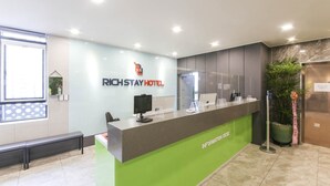A reception desk with a green base, a computer monitor, and a phone. The wall has a sign reading "RichStay Hotel".
