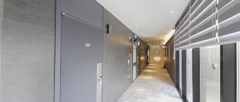 A modern hallway with a marble floor, grey walls, and a glass elevator on the right.