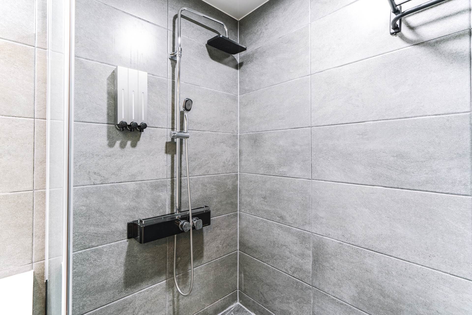 Un espace douche équipé d’un pommeau de douche à main, d’un panneau de commande fixé au mur et d’un motif de carrelage décoratif dans un coin.
