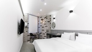 A modern hotel room with a large bed, a desk with a chair, a flat-screen TV mounted on the wall, and a small seating area with a cushioned chair and a whiteboard.