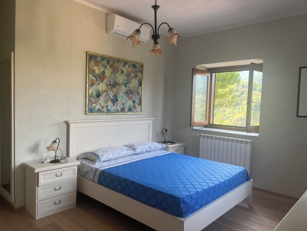 A bedroom with a bed, bedside tables, a radiator, a window with a view, and a painting on the wall.