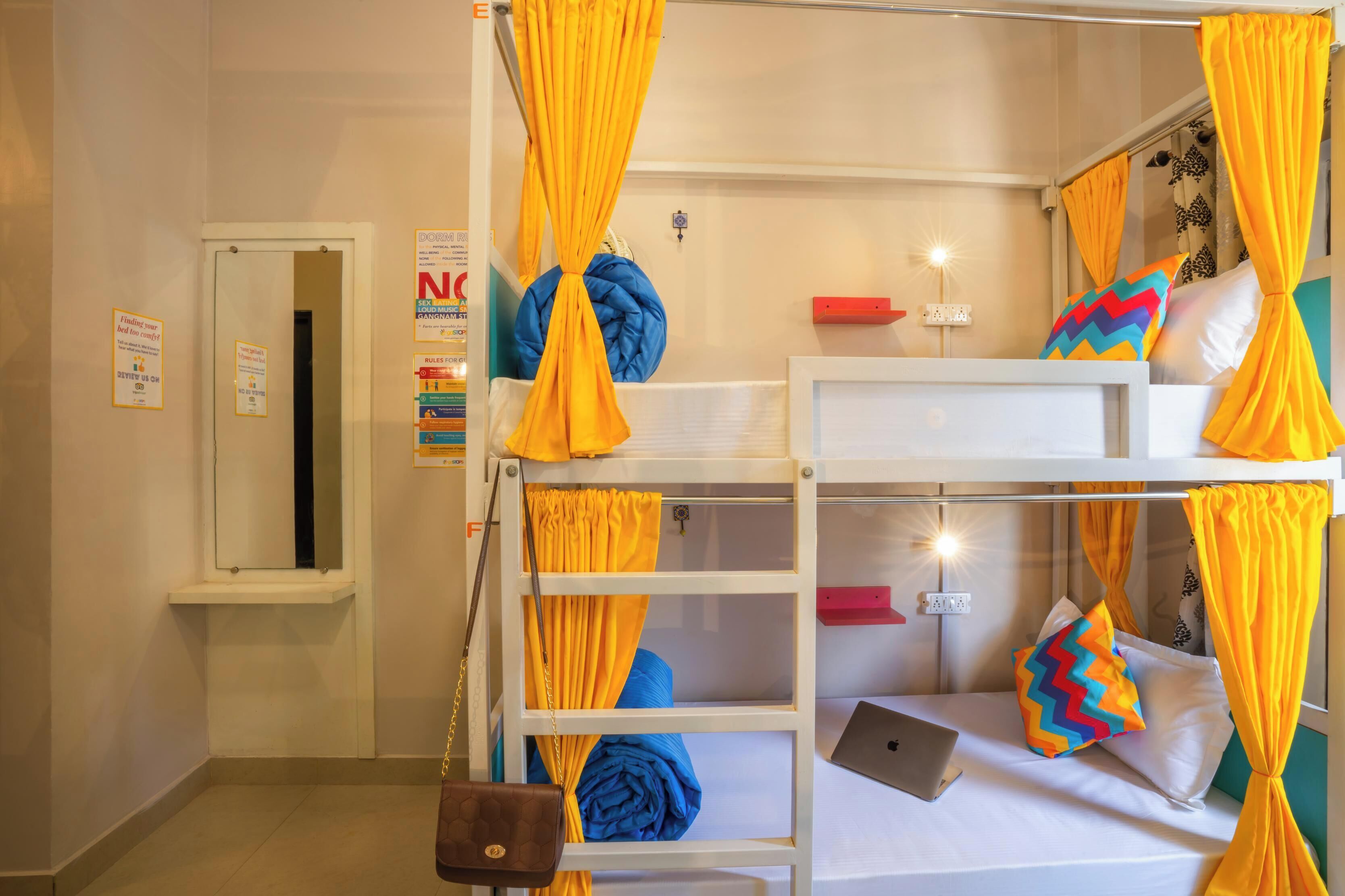 Habitación con literas, cortinas amarillas, una laptop en la camilla inferior y una pequeña estantería con un objeto rojo.