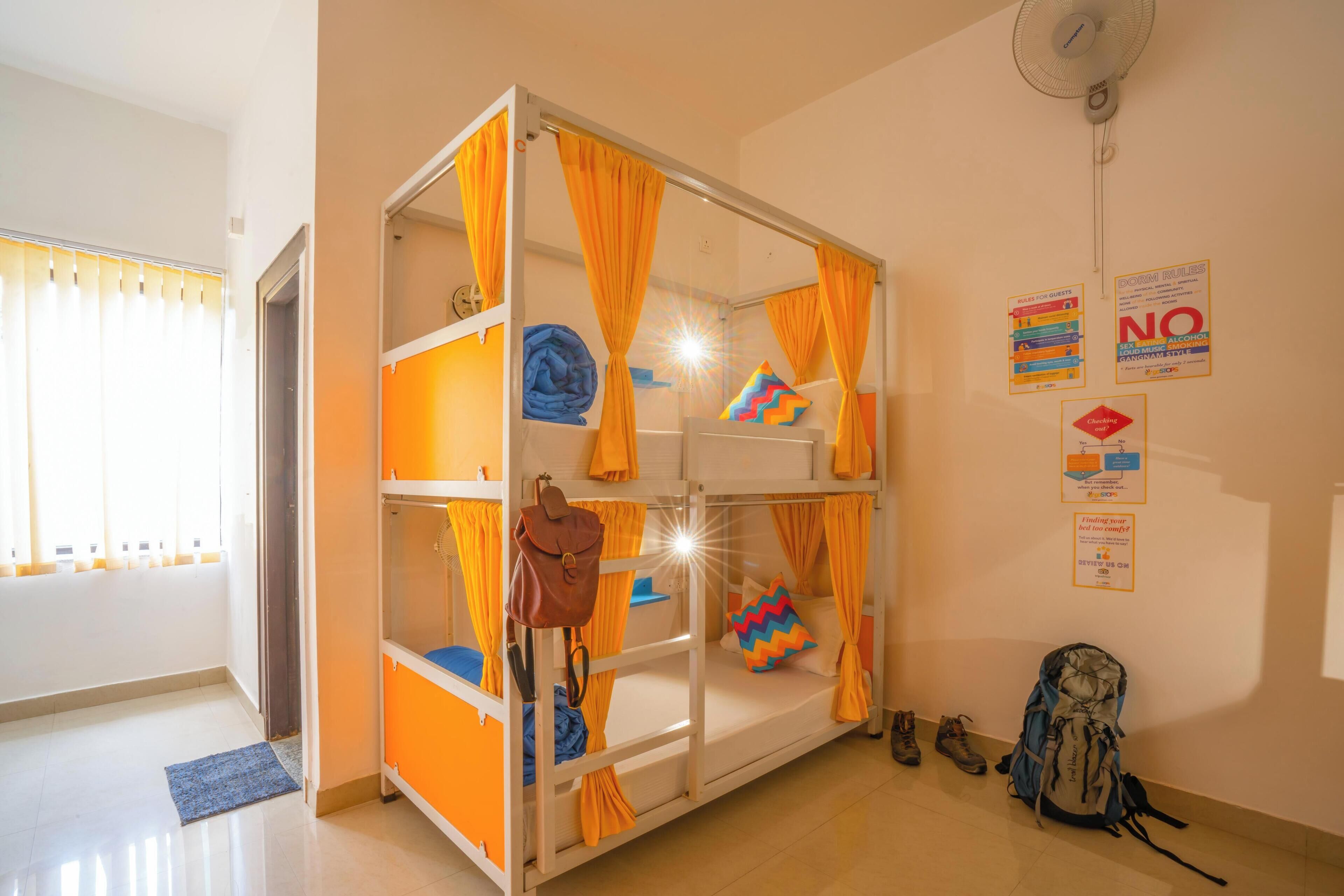 Habitación con literas, cortinas naranjas, un ventilador y un cartel con normas colgadas en la pared.