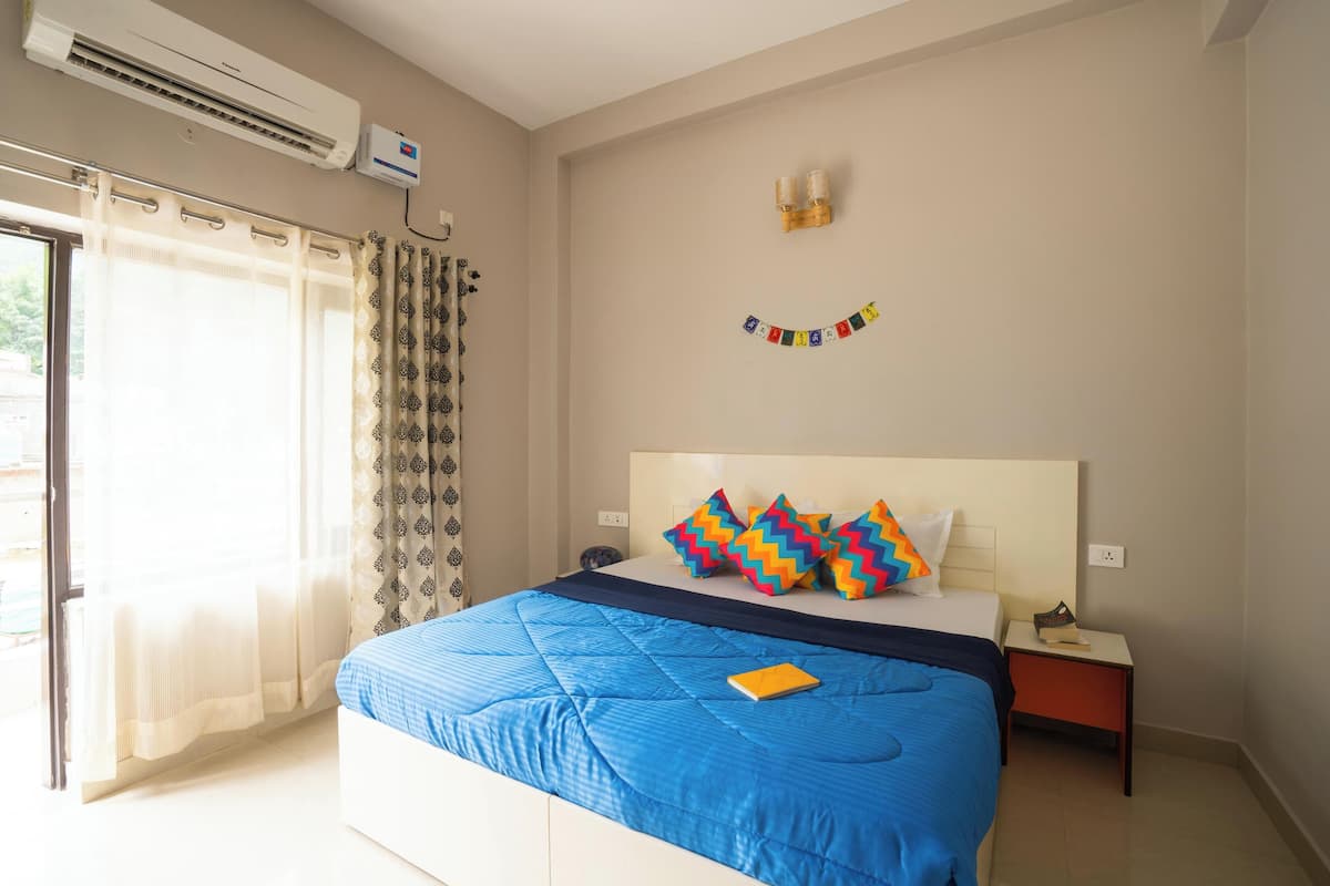 Un dormitorio con cama, mesita de noche, lámpara, ventana con cortinas y equipo de aire acondicionado.