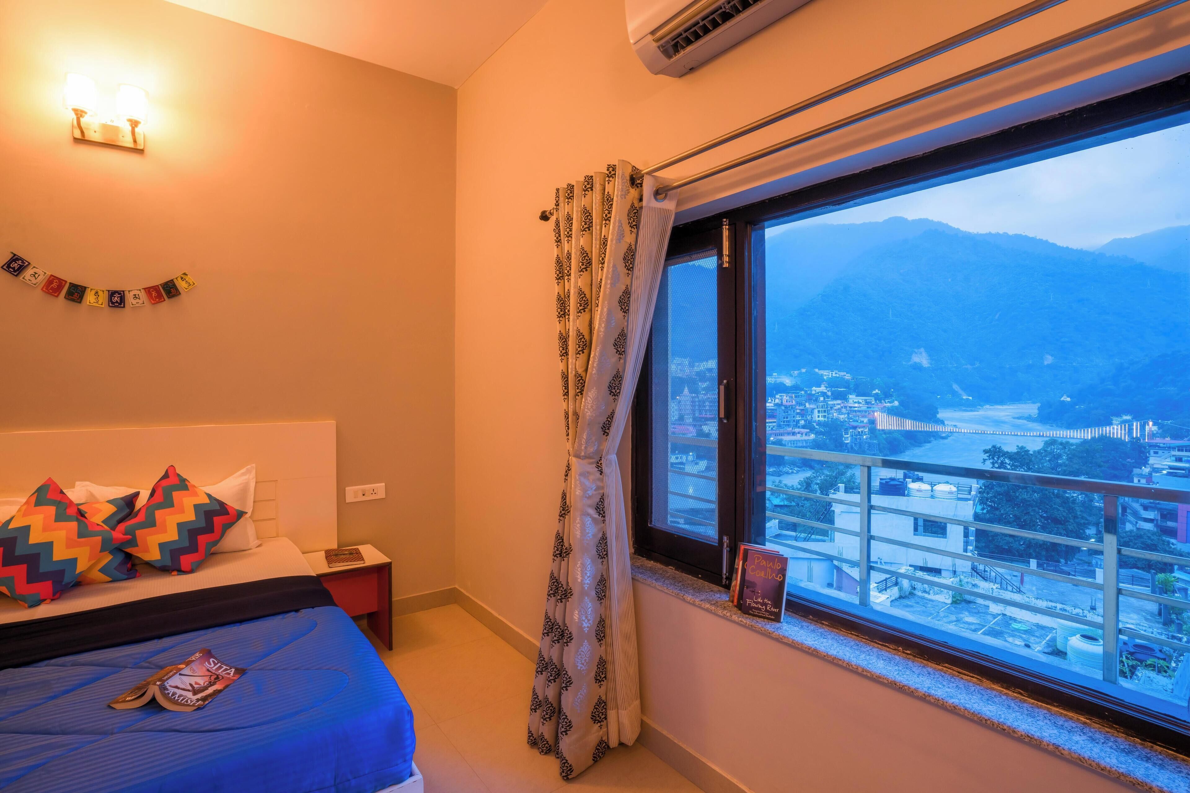 Una camera d'albergo con un letto, un comodino, una finestra vista città e montagne, e una tenda.