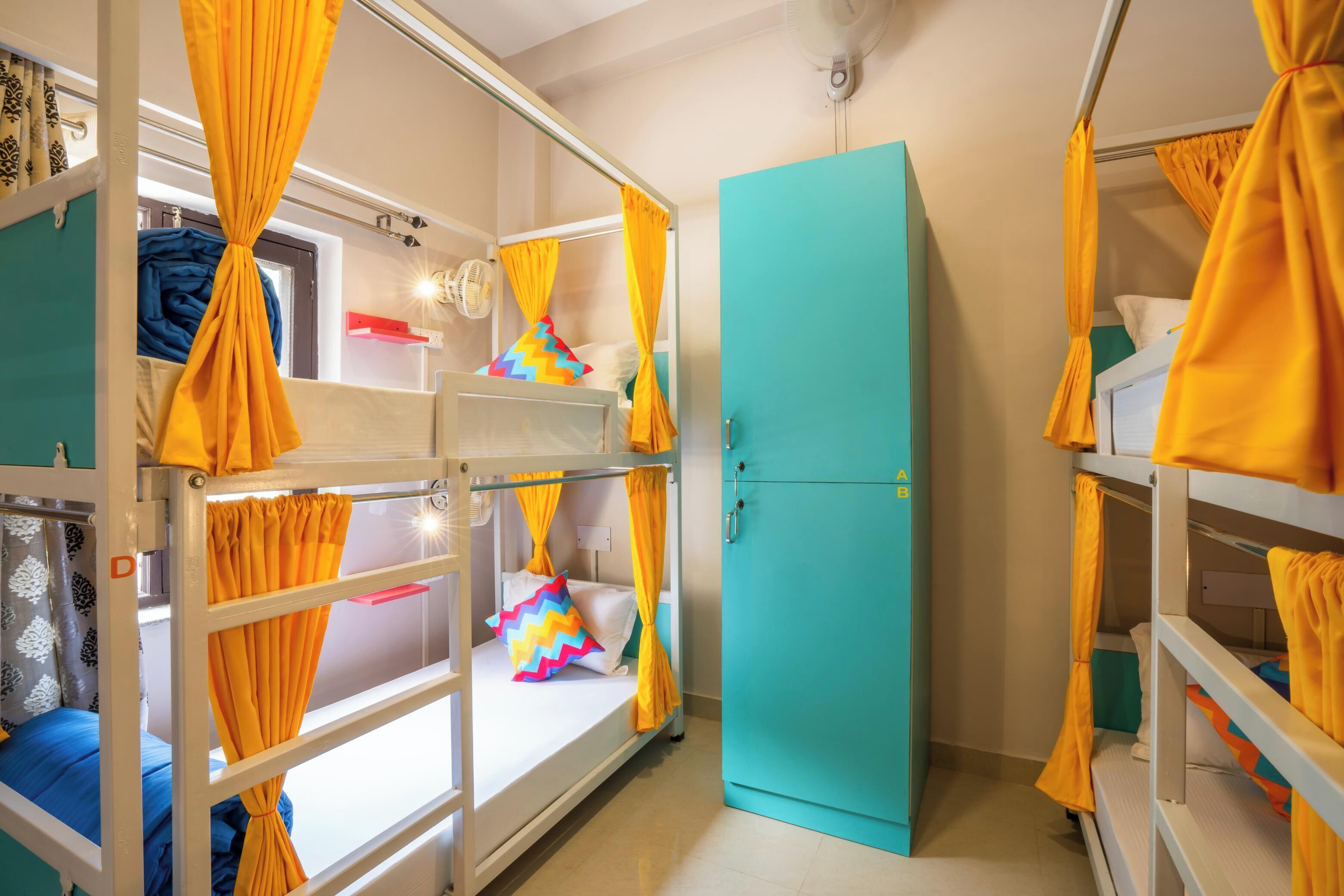 A dormitory room with bunk beds, a turquoise wardrobe, and yellow curtains.