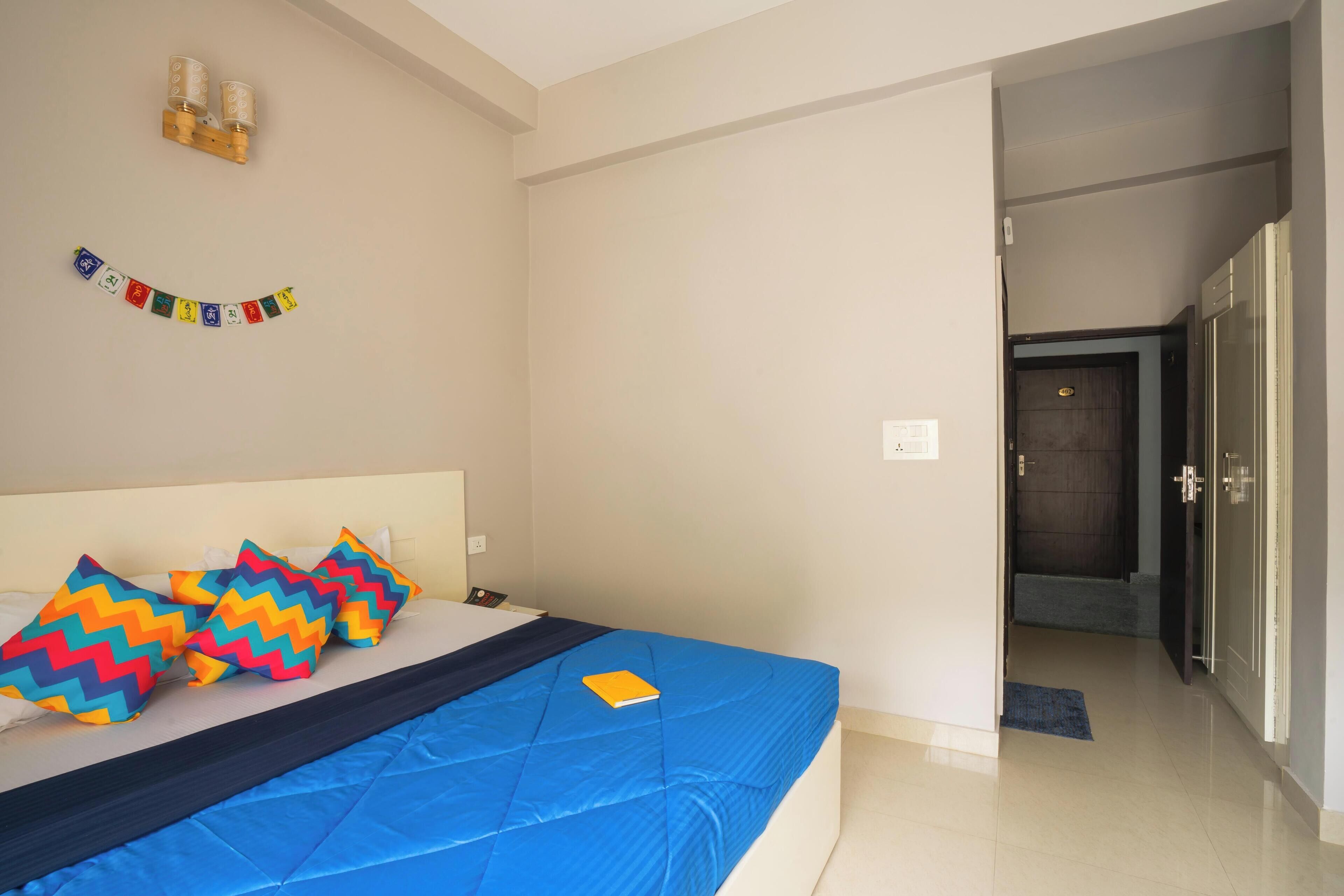 A hotel room with a bed, pillows, a wall-mounted light fixture, and a door to the right.