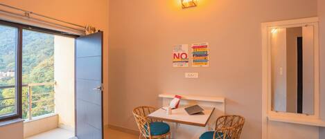 A hotel room with a large window, a desk with a chair, a wall-mounted light, and a poster with room rules.