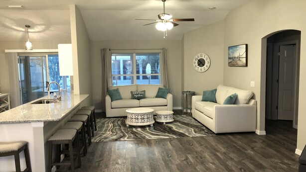 A modern kitchen with a central island, a dining area with bar stools, a living room with a sofa and coffee table, and a hallway leading to a door. - New Renovated House in Spanish Springs The Villages  (The Villages)