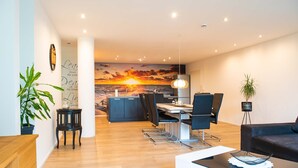 Un salon moderne avec un plancher en bois, un canapé noir, une table basse en verre et un coin repas offrant une vue sur le coucher de soleil sur l’océan.