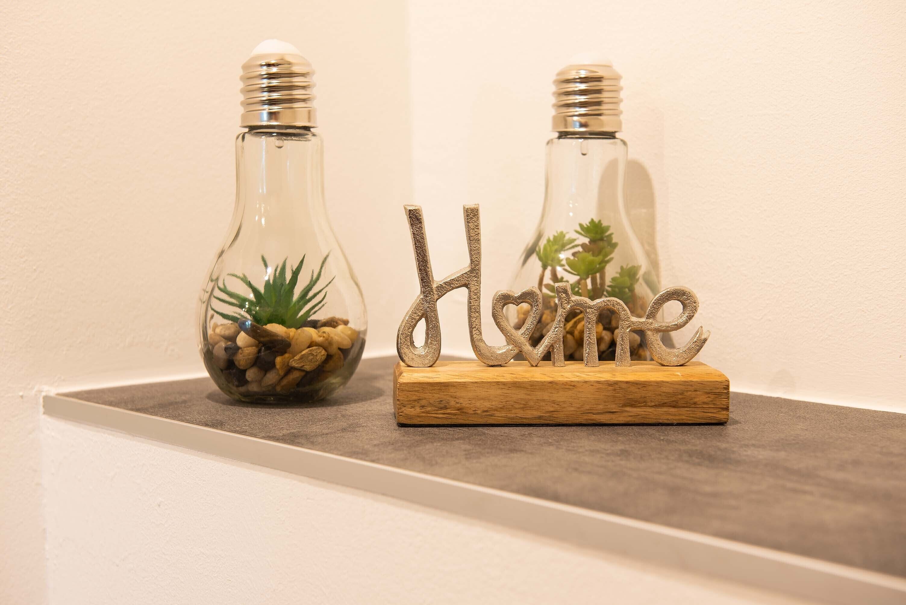 Deux bouteilles en verre contenant des plantes et des pierres, et une plaque en bois sur laquelle est inscrit le mot « Accueil », le tout placé sur une étagère.